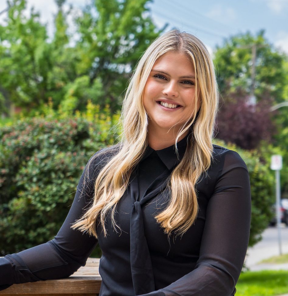 Chelsea Angus, wearing long blonde hair smiles while sitting outdoors on a sunny day. She is wearing a black blouse with a tie-style neckline. Behind her are green trees and shrubs, softly blurred in the background.