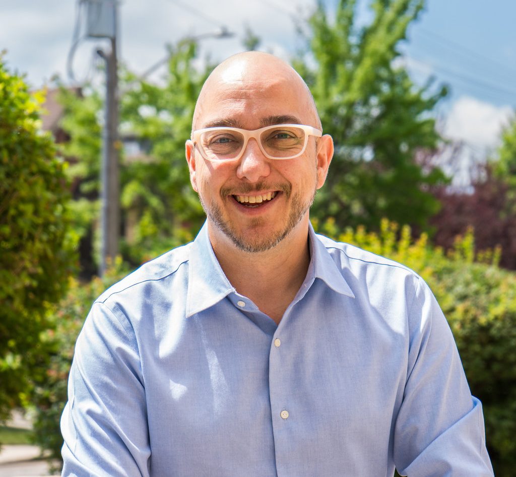 Nick Fundytus, wearing a short beard and light skin sits outdoors, smiling warmly. He is wearing light-coloured glasses and a pale blue button-up shirt. Behind him are leafy green trees and shrubs, softly blurred in the sunlight.